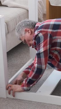 Evdeki bir masayı tornavidayla tamir eden bir adam, yerde oturuyor. Ev işi, mobilya, ve bir evde hizmet. Dikey video.