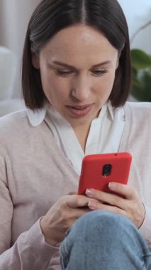 Evde akıllı telefonla çevrimiçi boş zamanlarını geçirirken gülen olumlu bir kadın. İletişim, internet gezintisi ve eğlencenin tadını çıkarırken aynı zamanda evde dinleniyor. Dikey video.