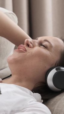 Gülümseyen kadın evde boş vakitlerin tadını çıkarıyor, kanepede dinlenirken kulaklıkla müzik dinliyor. Memnuniyet ve zevk, tam anlamıyla favori melodilerin ritmine dalmış. Dikey video.