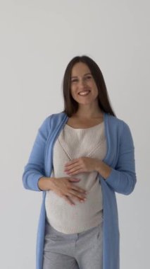 Beyaz bir arka planda duran şık ve iyimser hamile bir kadının portresi, gelecekteki anneliğin sevincini ve hamilelik sırasındaki sevgisini ifade ediyor. Dikey video.
