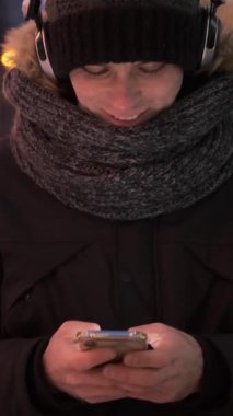 Neşeli adam internette boş zaman geçirmek için telefon kullanır ve kışın dışarıda dolaşırken müzik dinler, eğlenceli ve rahat bir gezintiden zevk alır. Dikey video.