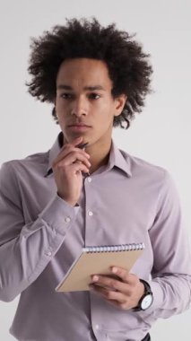 Düşünceli Afro-Amerikan iş adamı beyaz bir arka planda dururken not defterine not alıyor. Çalışmaya, ders çalışmaya ya da planlamaya odaklanmış. Dikey video.