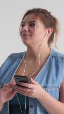 Dost canlısı bir kadın, beyaz bir zemin üzerinde telefon tutarken müzik dinler, şarkı söyler ve dans eder. Dinamik enerji ve neşe ifade eder. Dikey video.