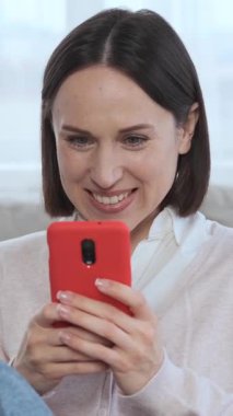 İnternet üzerinden sohbet eden bir kadın, evde boş zaman ve iletişim için telefon kullanıyor, internet gezintisinden zevk alıyor ve evde dinlenirken bağlı kalıyor. Dikey video.