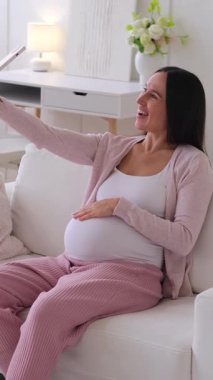 Orta yaşlı, Kafkasyalı hamile bir kadın arkadaşlarıyla evde cep telefonu kullanarak çevrimiçi video görüşmesine katılmış. Dijital iletişim konsepti. Dikey video.
