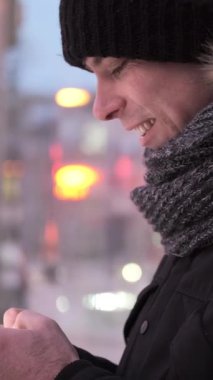 Adam kış şehrinde yürüyor, cep telefonuyla internetten mesaj atıyor. Açık hava aktivitesi ve dijital etkileşim konsepti. Dikey video.