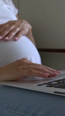 Hamile bir kadının evde karnını okşarken interneti karıştırmak için dizüstü bilgisayar kullandığı bir fotoğraf. Teknoloji ve boş zamanlarımızda evde olmanın rahatlığı. Dikey video.