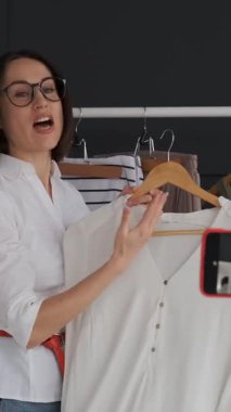 Heyecanlı bir kadın moda blogu için gardıroptan gelen bir telefonla canlı yayın yapıyor, kıyafet fikirlerini paylaşıyor, sosyal medya izleyicileriyle iletişim kuruyor ve çevrimiçi moda içeriği yaratıyor. Dikey video.
