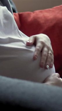 Sakin bir kadın evdeki koltukta karnını nazikçe okşayarak bebeği beklerken beklenti, sevgi ve özen gösteriyor. Dikey video.