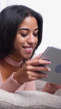 Gülümseyen Afro-Amerikalı kadın evde tablet bilgisayar kullanarak internet üzerinden mesajlaşıyor, rahat bir ortamda internet eğlencesinin ve eğlencenin tadını çıkarıyor. Dikey video.