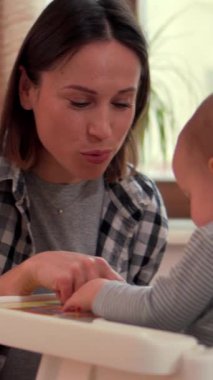 Mutlu anne, bebek bebekle birlikte mutfaktaki masada öğreniyor, neşeli ve eğitici bir anı paylaşıyor. Dikey video.