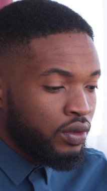 Ciddi bir Afro-Amerikan serbest çalışanı evde dizüstü bilgisayarda çalışıyor, iş, muhasebe ve iletişim işleriyle uğraşıyor. Uzaktan kumandaya odaklan ve kendini işine ver. Dikey video.