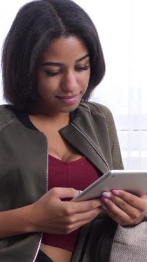 Gülümseyen Afro-Amerikalı kadın online eğlence ve internet gezintisine çıktı. Bir tablet bilgisayarla evdeki kanepede dinleniyor. Dikey video.