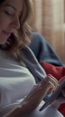 Mutlu hamile kadın boş zamanlarını evde eğlenmek ve rahatlamak için bir tablet bilgisayar kullanarak geçiriyor. Dikey video.