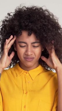 Mutsuz ve sağlıksız Afrikalı Amerikalı kadın beyaz bir arka planda baş ağrısı ya da migrenden muzdarip, umutsuzluk ve yorgunluk gösteriyor. Dikey video.