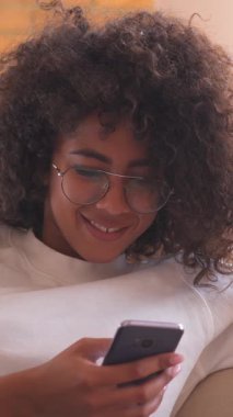 Mutlu Afro-Amerikalı kadın evde rahatlarken akıllı telefondan internet aktivitelerinin keyfini çıkarıyor, mesajlaşıyor ve boş zamanlarında rahatlıyor. Dikey video.