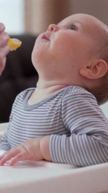 Gülümseyen küçük bebek mutfakta annesiyle birlikte yemek yiyor. Bakım, beslenme ve neşe dolu bir anı paylaşıyor. Dikey video.