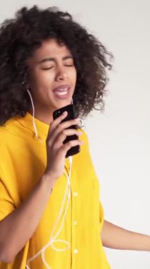 Neşeli Afrikalı Amerikalı kadın beyaz bir arka planda şarkı söylüyor, dans ediyor ve müzik dinliyor. Enerji, müzik ve pozitiflik. Dikey video.