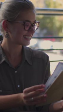 Gülen kadın kitap okuyor ve gündüzleri dışarıda eğleniyor. Rahatlama, keyif ve edebiyat aşkı. Dikey video.