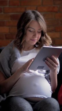 Gülümseyen hamile kadın evdeki kanepede dinleniyor, dijital boş zamanlarında online eğlence için tablet bilgisayar kullanıyor. Dikey video.