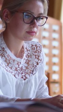 İnternette, bir kütüphanede dizüstü bilgisayarla çalışan düşünceli bir kadın, iletişim ve ağ kurmaya odaklanıyor. Geleneksel bir ortamda konsantrasyon ve modern teknoloji. Dikey video.