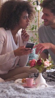 Aile çifti yatak odasında online eğlence için telefon kullanıyor. Evde huzurlu bir atmosferde birlikte sabah kahvaltısının tadını çıkarıyoruz. Dikey video.