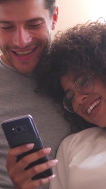 Heyecanlı kız arkadaş ve erkek arkadaş birlikte gülerken evde internet aktiviteleri ve eğlence için telefon kullanıyorlar. Dikey video.