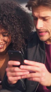 Mutlu çift açık havada telefonları kullanarak internette eğlenmeyi seviyor. Boş zaman, teknoloji ve paylaşılan mutluluk. Dikey video.