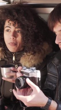 Neşeli erkek arkadaş ve kız arkadaş dışarıda fotoğraf çektirirken bir arabanın bagajında birlikte oturmaktan hoşlanırlar. Mutlu ifadeler, hatırlanmaya değer birliktelik anları. Dikey video.