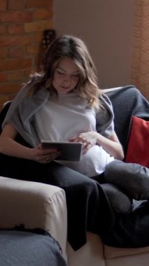 Gülümseyen hamile kadın evde tablet bilgisayar kullanarak internette geziniyor, boş zamanlarında rahatlık ve iletişimin tadını çıkarıyor. Dikey video.