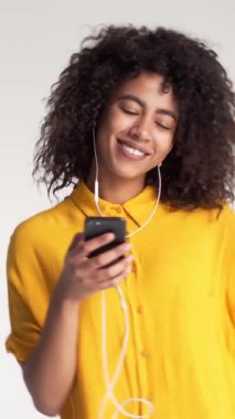 Neşeli ve tatmin olmuş Afrikalı Amerikalı kadın beyaz arka planda akıllı bir telefonla dans ediyor ve müzik dinliyor. Neşe, teknoloji ve modern yaşam tarzı. Dikey video.