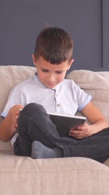 Çocuk evdeki kanepede dinleniyor, online eğlence ve eğlence için tablet kullanıyor. Siber uzayda rahatlama ve eğlence. Dikey video.