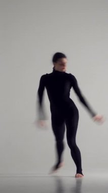Kadın, siyah bir sahne kostümüyle beyaz bir arka planda, ilham ve esneklik sergileyerek çağdaş bir dans sergiliyor. Performans, sanatçılık ve zarafet. Dikey video.