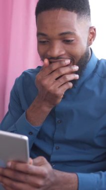 Heyecanlı bir Afro-Amerikan erkeği evde bir tabletle internetten eğlenmeyi, gülmeyi ve rahatlamayı seviyor. Heyecan, rahatlama ve eğlence. Dikey video.