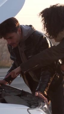 Endişeli eş ve koca, açık kapüşonlu kırık arabayı gezide araç sorunlarıyla karşı karşıya bırakarak inceliyor. Dikey video.