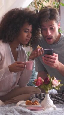 Karı ve koca sabah kahvaltısını yatak odasında yapıyorlar. Boş zamanlarında internet eğlencesi için hoş geldin atmosferi için telefon kullanmak. Dikey video.