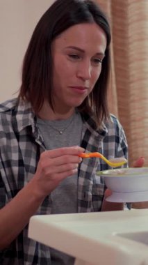 Gülümseyen anne, bebeğini evde öğle yemeği sırasında kaşıkla besliyor, yemek veriyor ve yemek yiyor. Dikey video.