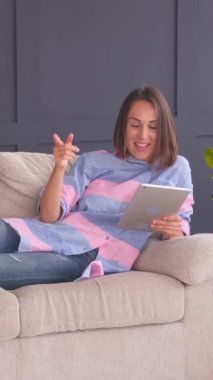 Evdeki kanepede uzanmış, elinde tabletle internette gezinen neşeli bir kadın. Boş zamanlarında rahatlama ve keyif. Dikey video.