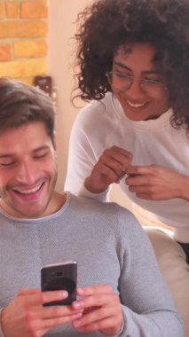 Keyifli kadın ve erkek boş zamanlarında evde dinleniyor, telefonu çeşitli online aktiviteler ve eğlence için mutlu bir şekilde kullanıyor. Dikey video.