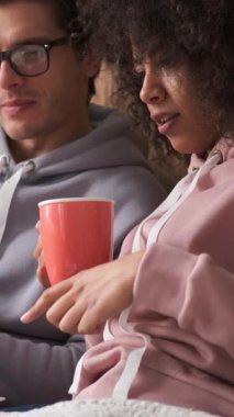 Kız arkadaş ve erkek arkadaş evde dizüstü bilgisayarla internet eğlencesi yaparken çay içiyorlar. Rahat bir dijital rahatlık. Dikey video.