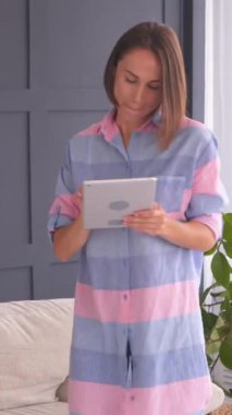 Dost canlısı bir kadın dans ediyor ve hafta sonu evde bir tabletle vakit geçirmekten hoşlanıyor. Boş zaman, rahatlama ve neşeli bir atmosfer. Dikey video.