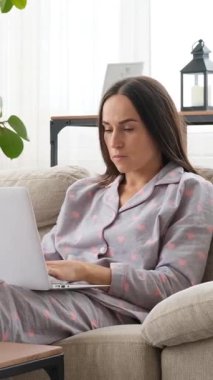 İnternette evdeki bir kanepede dizüstü bilgisayar kullanarak çalışan ciddi bir kadın, rahat bir iş ortamında odaklanarak ve adanmışlıkla internete bağlanıyor. Dikey video.