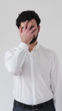 Adam beyaz bir arkaplanda utancı, sıkıntıyı, umutsuzluğu ve hayal kırıklığını açık ve anlaşılır bir şekilde ifade eden bir facepalm hareketi yapıyor. Dikey video.