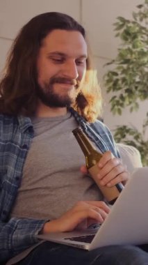 Hevesli bir adam evde bira yudumlarken internette sohbet ediyor, boş zamanlarında dinleniyor ve sevinçle dijital etkileşime giriyor. Dikey video.