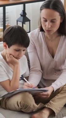 Bakıcı bir anne oğluna ev ödevlerinde ve egzersizlerinde yardım ediyor, sıcak ve eğitici bir ortamda okul derslerini destekliyor. Dikey video.