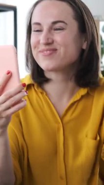 Dost canlısı bir kadın internet eğlencesi için akıllı telefon kullanıyor, evde boş zamanlarında dans ediyor, hareketli ve çekici bir atmosferde rahatlamayı kucaklıyor. Dikey video.