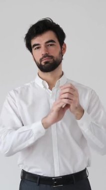 Tatmin olmuş bir adam beyaz bir arkaplanda el çırpıyor. Basit bir ortamda keyif, saygı ve sıcak bir selamlama sergiliyor. Dikey video.