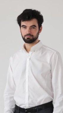 Gülümseyen ve şakacı bir adam, beyaz bir arkaplanda macera çağrıştıran ve karizmatik ve çekici bir tavırla flört eden bir daveti işaret eder. Dikey video.