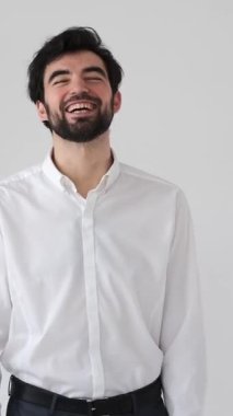 Beyaz bir arkaplana karşı bir portrede gülen olumlu adam sıcak ve çekici bir ifadeyle şakadan neşe, eğlence ve neşe saçıyor. Dikey video.