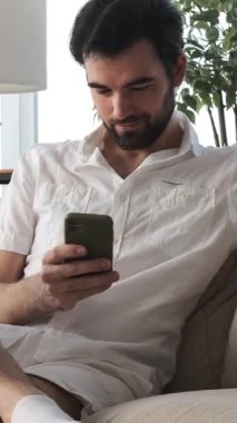 Ciddi bir adam, evde online boş zaman ve mesajlaşma için telefon kullanıyor, rahat bir iç mekanda dijital iletişimle eğleniyor. Dikey video.